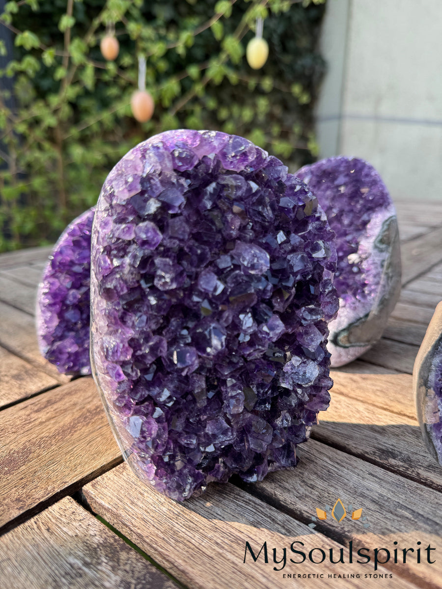 Ein Nahaufnahme von drei glänzenden Amethyst-Drusen, die auf einer hölzernen Tischplatte vor einem verschwommenen grünen Hintergrund mit hängenden Dekorationen stehen.